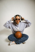 Person mit Sonnenbrille und Kopfhörern, die einen Basketball hält, trägt einen oversized Unisex-Hoodie in Hellblau mit Drop-Shoulder-Passform. Der Hoodie mit doppelten Nähten und aus schwerer French Terry Baumwolle bietet Komfort, Langlebigkeit und Atmungsaktivität. Auf der Brust steht der Schriftzug 'don't let your past keep you'. Der zweite Teil der Botschaft, 'from your best', ist nicht sichtbar. Das Logo der Marke 'BAKLAVA' ist ebenfalls zu erkennen. Inspiriert von Kendrick Lamar’s Song 'Die Hard'.